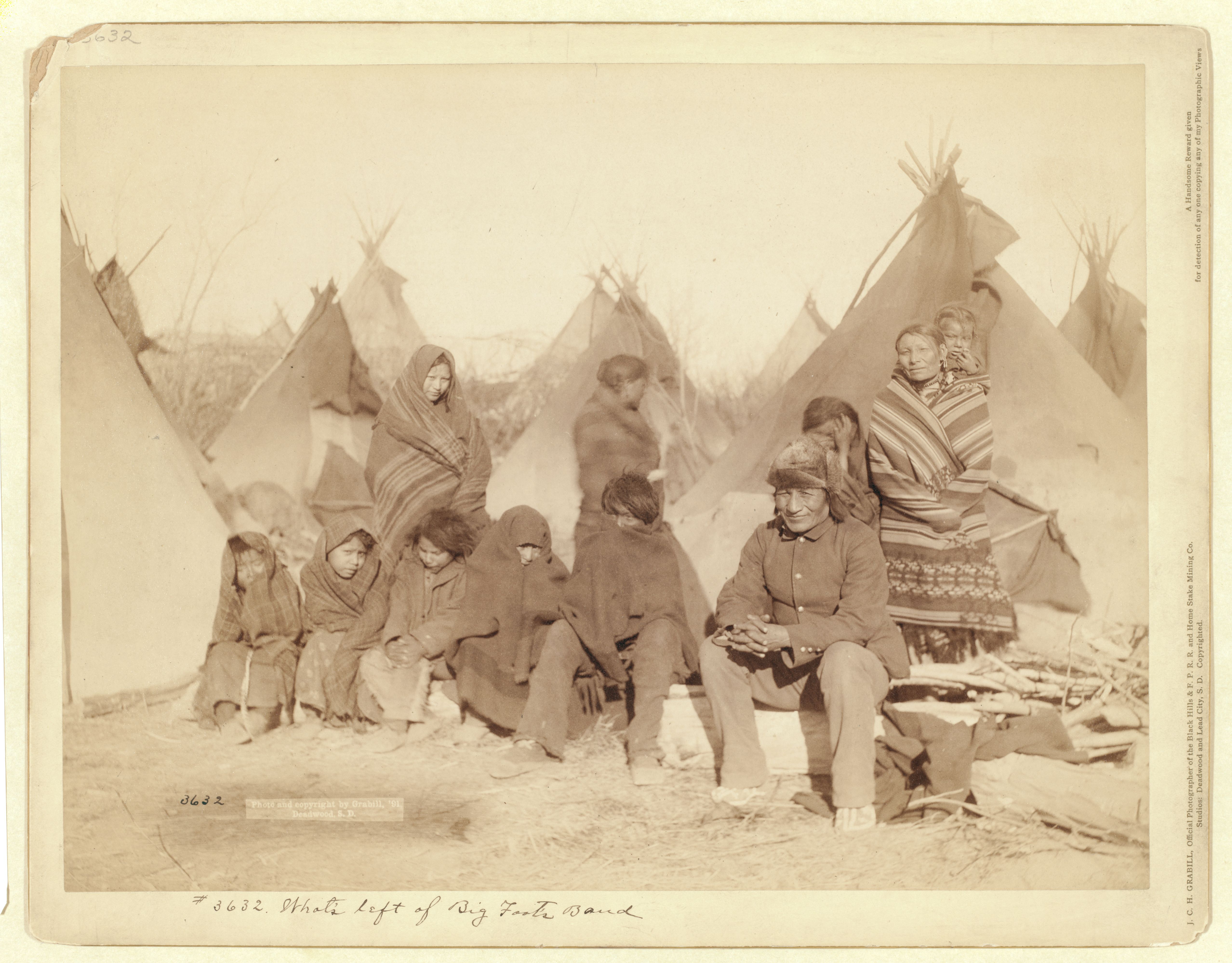 Historic, sepia photograph of eleven Plains Indians, including men, women, and children sitting and standing in front of eight tipis.