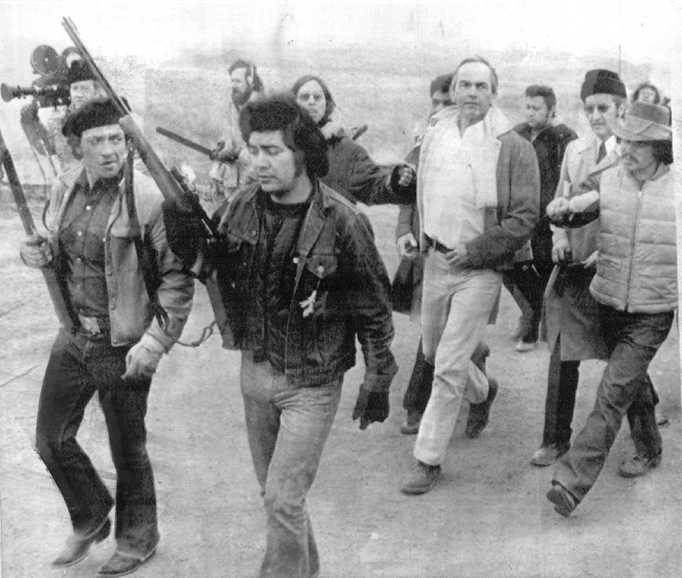 Black and white photograph of ten men walking. Some are armed. One man is carrying a large camera.