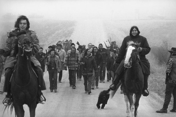 Black and white photograph of two Native people on horseback leading a group of AIM protestors on a dirt road in Pine Ridge South Dakota. All are facing the camera. Some of the protestors are carrying rifles.