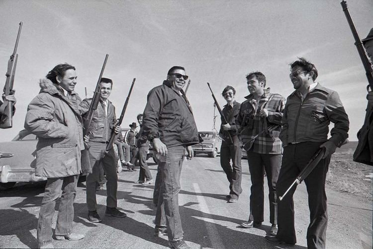 Black and white photograph of Dick Wilson standing in the middle of a road surrounded by ten Native men who are holding rifles over their shoulders and smiling.