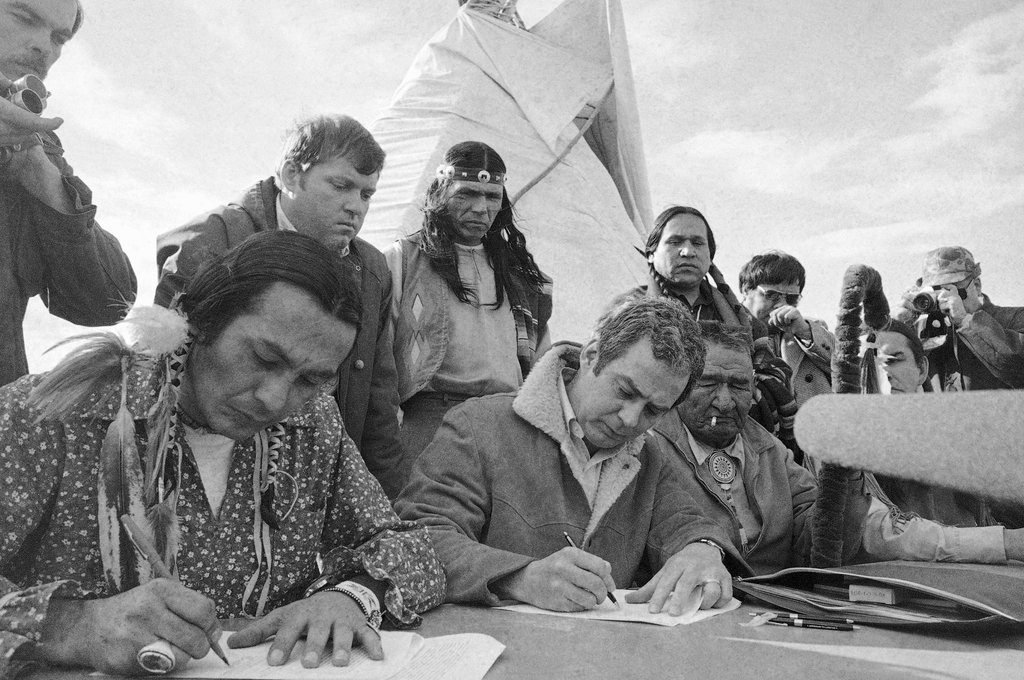 Black and white photograph of Russell Means (front left) signing a settlement with assistant Attorney General Kent Frizzell in Wounded Knee, South Dakota. Six Native and White men, including Dennis Banks wearing a headband, stand behind Means and Frizzell. This event is taking place outside and there is a tipi in the background.