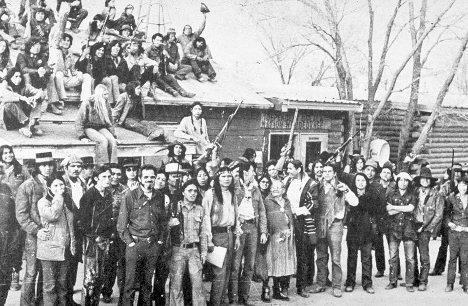 Black and white photograph of a large crowd of Native men and women standing in front of and sitting on top of a building in Wounded Knee, South Dakota. The text ”Independent Oglala Nation Wounded Knee” appears on the door of the building.