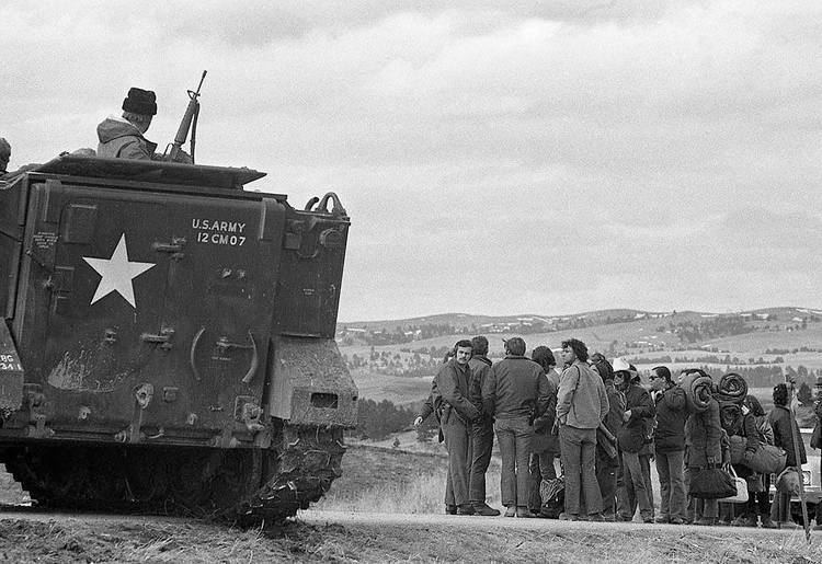 Black and white photograph of a U.S. Army tank blocking the road. An armed man emerges from the top of the tank and a small crowd of people holding sleeping bags and duffle bags stand on the road downhill from the tank.