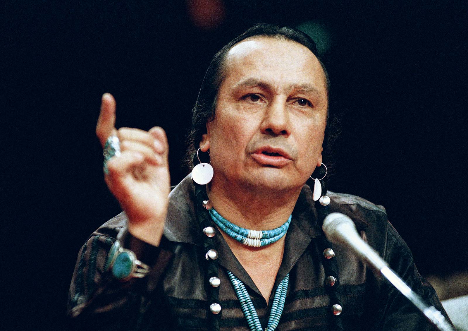 Color photograph of Russell Means wearing turquoise jewelry and speaking into a microphone.