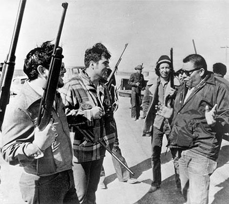 Black and white photograph of Dick Wilson, right, talking to three Native men holding rifles.