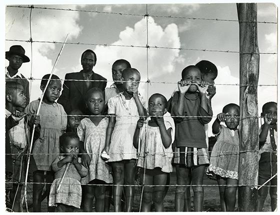 A group of children and a few men gaze from behind a barbed-wire fence that marks the boundary of the Moroka township in Soweto, Johannesburg, South Africa